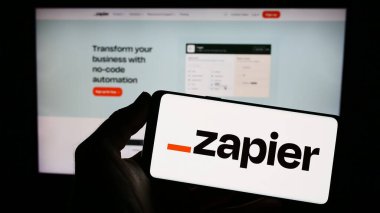 Stuttgart, Germany - 03-30-2023: Person holding smartphone with logo of US automation software company Zapier Inc. on screen in front of website. Focus on phone display. clipart