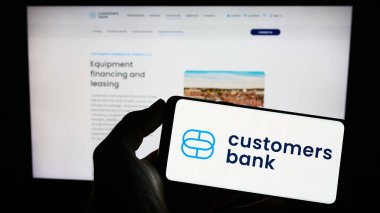 Stuttgart, Almanya - 04-01-2023: ABD 'li finans şirketi Customers Bancorp Inc.' in logosuyla cep telefonu sahibi kişi iş sayfasının önünde ekranda. Telefon ekranına odaklan.