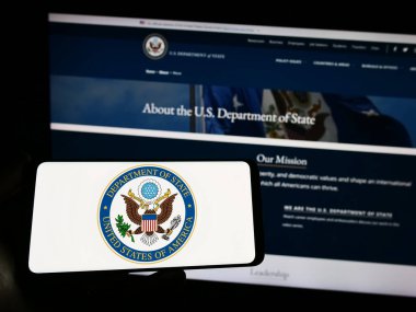 Stuttgart, Germany - 04-01-2023: Person holding smartphone with seal of United States Department of State (DOS) on screen in front of website. Focus on phone display. clipart