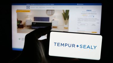 Stuttgart, Germany - 01-15-2025: Person holding mobile phone with logo of US bedding company Tempur Sealy International Inc. on screen in front of web page. Focus on phone display. clipart