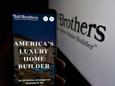 Stuttgart, Germany - 01-15-2025: Person holding cellphone with webpage of US homebuilding company Toll Brothers Inc. on screen in front of logo. Focus on center of phone display. clipart