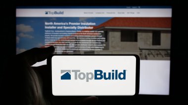 Stuttgart, Almanya - 01-15-2025: Üzerinde ABD 'li inşaat malzemeleri şirketi TopBuild Corp' un logosu olan cep telefonu sahibi kişi iş sayfasının önünde ekranda görülüyor. Telefon ekranına odaklan.