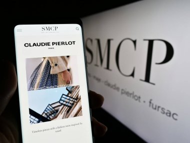 Stuttgart, Germany - 03-25-2023: Person holding cellphone with webpage of French luxury fashion company SMCP S.A. on screen in front of logo. Focus on center of phone display. clipart