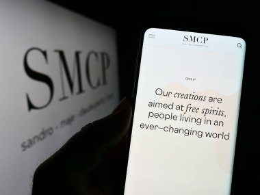 Stuttgart, Almanya - 03-25-2023: Fransız lüks moda şirketi SMCP S.A. 'nın web sayfası ile akıllı telefon sahibi kişi logonun önünde ekranda. Telefon ekranının merkezine odaklan.