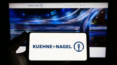 Stuttgart, Germany - 03-25-2023: Person holding smartphone with logo of logistics company Kuehne Nagel International AG on screen in front of website. Focus on phone display. clipart