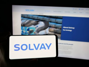 Stuttgart, Germany - 01-24-2025: Person holding smartphone with logo of Belgian chemical company Solvay S.A. on screen in front of website. Focus on phone display. clipart