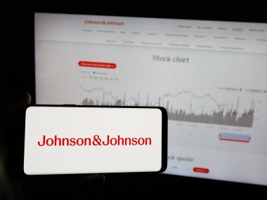 Stuttgart, Germany - 01-30-2025: Person holding smartphone with logo of US pharmaceutical company Johnson and Johnson on screen in front of website. Focus on phone display. clipart