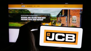 Stuttgart, Almanya - 03-04-2025: İngiliz J.C. Bamford Excavators Limited (JCB) şirketinin logosuna sahip cep telefonu sahibi kişi iş sayfasının önünde ekranda görülüyor. Telefon ekranına odaklan.