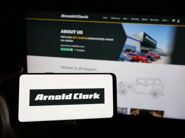 Stuttgart, Almanya - 03-04-2025: Araba satıcısı şirketi Arnold Clark Automobiles Ltd. 'nin logosunu taşıyan kişi iş sitesinin önünde ekranda. Telefon ekranına odaklan.