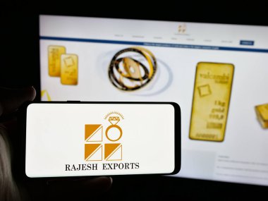 Stuttgart, Almanya - 03-13-2025: Hintli altın şirketi Rajesh Exports Limited 'in logosuna sahip akıllı telefon sahibi kişi iş sitesinin önünde ekranda görülüyor. Telefon ekranına odaklan.