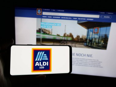 Stuttgart, Almanya - 03-13-2025: Alman perakende şirketi ALDI 'nin logosuna sahip akıllı telefon sahibi kişi iş sitesinin önünde ekrana yansıdı. Telefon ekranına odaklan.