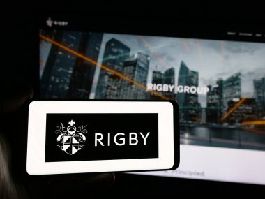 Stuttgart, Almanya - 03-13-2025: İngiliz firması Rigby Group plc 'in logosunu taşıyan kişi iş sitesinin önünde ekranda. Telefon ekranına odaklan.