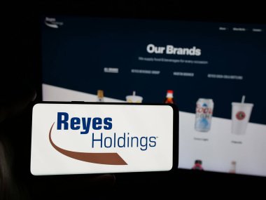 Stuttgart, Almanya - 03-06-2025: ABD 'li gıda ve içecek şirketi Reyes Holding LLLC' nin logosuna sahip akıllı telefon sahibi kişi iş sitesinin önünde ekranda görülüyor. Telefon ekranına odaklan.