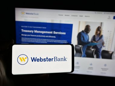 Stuttgart, Almanya - 03-07-2025: ABD 'li finans şirketi Webster Financial Corporation' ın logosuyla akıllı telefon sahibi kişi, iş dünyasının web sitesi önünde ekranda. Telefon ekranına odaklan.