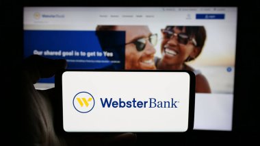 Stuttgart, Almanya - 03-07-2025: ABD 'li finans şirketi Webster Financial Corporation' ın logosuyla cep telefonu sahibi kişi iş sayfası önünde ekranda. Telefon ekranına odaklan.