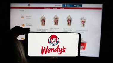 Stuttgart, Almanya - 03-07-2025: ABD 'li restoran şirketi Wendy' nin Uluslararası LLC 'sinin logosuyla cep telefonu sahibi kişi iş sayfasının önünde ekranda. Telefon ekranına odaklan.