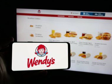 Stuttgart, Almanya - 03-07-2025: ABD 'li restoran şirketi Wendy' nin Uluslararası LLC 'sinin logosunu taşıyan kişi iş sitesinin önünde ekranda. Telefon ekranına odaklan.