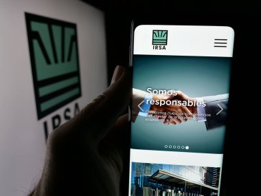 Stuttgart, Almanya - 03-31-2025: Logonun önündeki ekranda Inversiones y Temsilcileri S.A. (IRSA) logosunun logosu bulunan kişi. Telefon ekranının merkezine odaklan.
