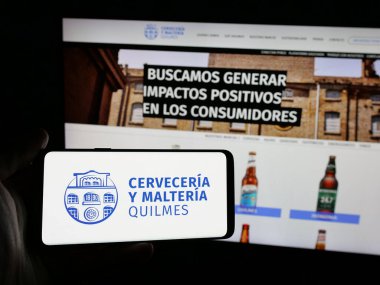 Stuttgart, Almanya - 04-01-2025: Web sitesi önünde bira üreten Cerveceria y Malteria Quilmes şirketinin logosuna sahip akıllı telefon sahibi kişi. Telefon ekranına odaklan.