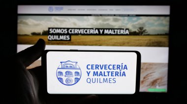 Stuttgart, Almanya - 04-01-2025: Web sayfası önünde bira üreten Cerveceria y Malteria Quilmes şirketinin logosuyla cep telefonu tutan kişi. Telefon ekranına odaklan.