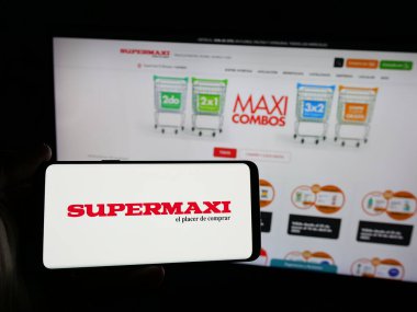 Stuttgart, Almanya - 04-09-2025: Ekvador perakende şirketi Supermaxi 'nin logosuna sahip akıllı telefon sahibi kişi web sitesinin önünde ekranda. Telefon ekranına odaklan.