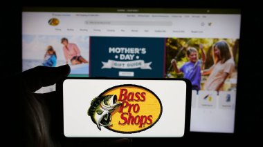 Stuttgart, Almanya - 05-02-2025: Web sayfası önünde üzerinde ABD 'li Bass Pro Shops (BPS Direct LLC) şirketinin logosu bulunan cep telefonu sahibi kişi. Telefon ekranına odaklan.