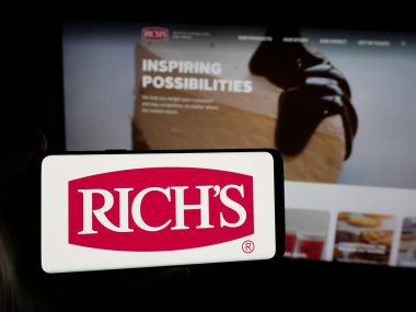 Stuttgart, Almanya - 05-15-2025: Rich Products Corporation (Rich 's) şirketinin logosunu elinde tutan kişi web sitesinin önünde ekranda. Telefon ekranına odaklan.