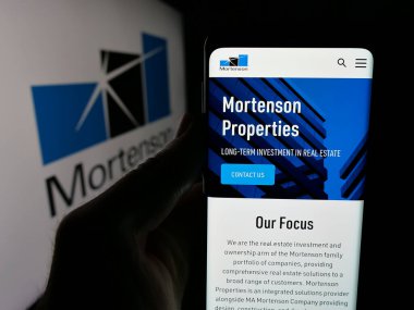 Stuttgart, Almanya - 05-17-2025: İnşaat işletmeleri M.A. web sayfası ile akıllı telefon tutan kişi Mortenson Şirketi logonun önünde ekranda. Telefon ekranının merkezine odaklan.