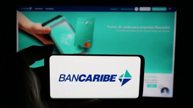 Stuttgart, Almanya - 05-18-2025: Banco del Caribe C.A. şirketinin logosuyla cep telefonu tutan kişi (Bancaribe) Web sayfasının önünde ekranda. Telefon ekranına odaklan.