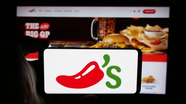 Stuttgart, Almanya - 05-25-2025: İnternet sayfasının önünde ABD 'li restoran zinciri şirketi Chilis Grill Bar' ın logosuyla cep telefonu tutan kişi. Telefon ekranına odaklan.