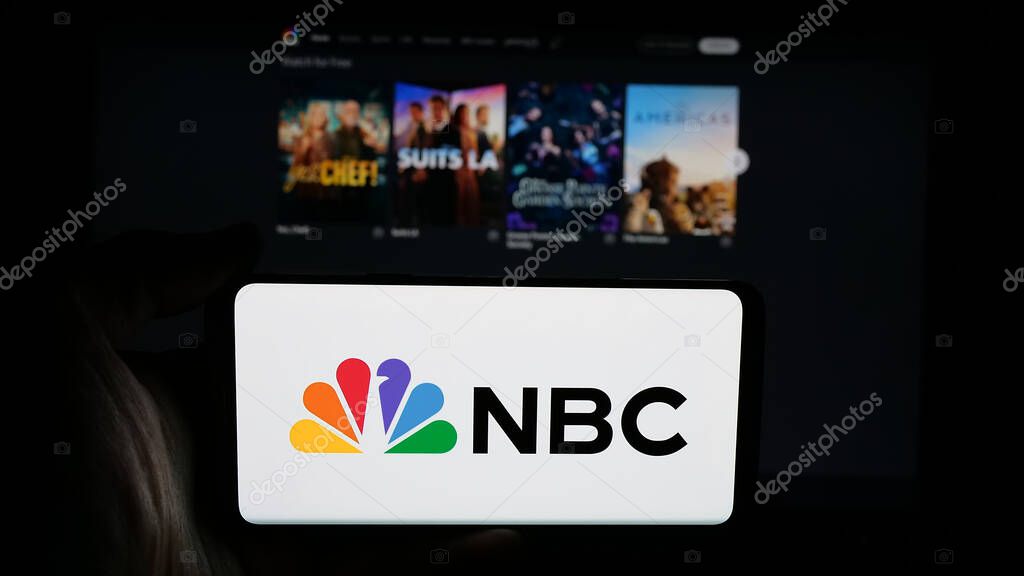 Stuttgart, Germany - 05-25-2025: Person holding cellphone with logo of network National Broadcasting Company (NBC) on screen in front of webpage. Focus on phone display.