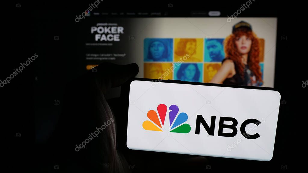 Stuttgart, Germany - 05-25-2025: Person holding mobile phone with logo of network National Broadcasting Company (NBC) on screen in front of web page. Focus on phone display.