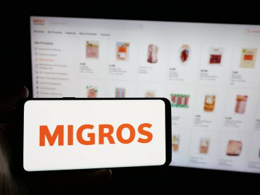 Stuttgart, Almanya - 05-25-2025: İsviçreli süpermarket şirketi Migros 'un logosunu elinde tutan kişi web sitesinin önünde ekranda. Telefon ekranına odaklan.