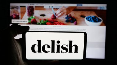 Stuttgart, Almanya - 05-27-2025: İnternet sayfası önünde ABD gıda çevrimiçi dergisi Delish.com (Hearst) logosu olan cep telefonu sahibi kişi. Telefon ekranına odaklan.
