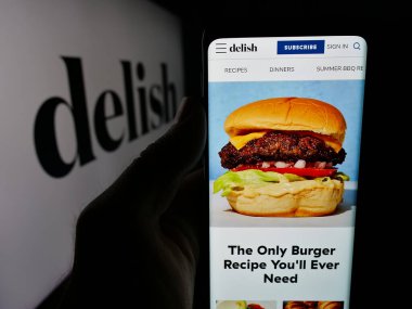 Stuttgart, Almanya - 05-27-2025: ABD gıda çevrimiçi dergisi Delish.com 'un (Hearst) internet sayfası logonun önünde cep telefonu tutan kişi. Telefon ekranının merkezine odaklan.