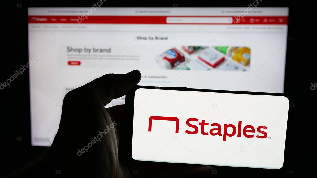 Stuttgart, Germany - 05-26-2025: Person holding mobile phone with logo of US office supply retail company Staples Inc. on screen in front of web page. Focus on phone display.