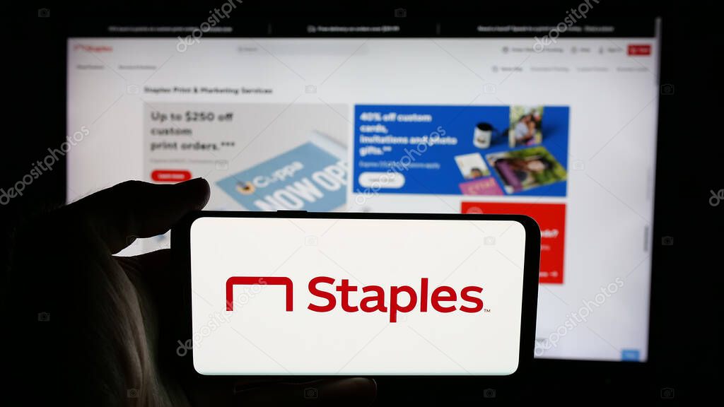 Stuttgart, Germany - 05-26-2025: Person holding cellphone with logo of US office supply retail company Staples Inc. on screen in front of webpage. Focus on phone display.