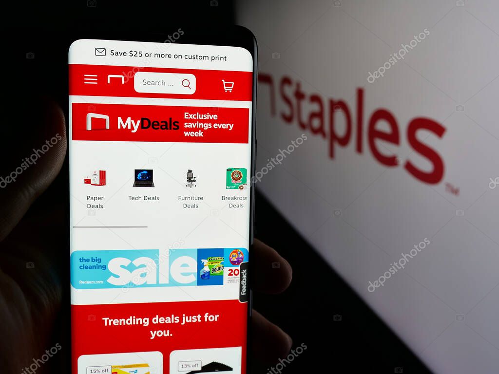Stuttgart, Germany - 05-26-2025: Person holding smartphone with web page of US office supply retail company Staples Inc. on screen in front of logo. Focus on center of phone display.