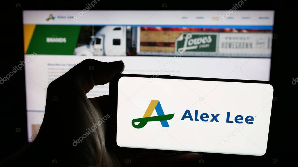 Stuttgart, Germany - 06-05-2025: Person holding mobile phone with logo of US grocery wholesale company Alex Lee Inc. on screen in front of web page. Focus on phone display.