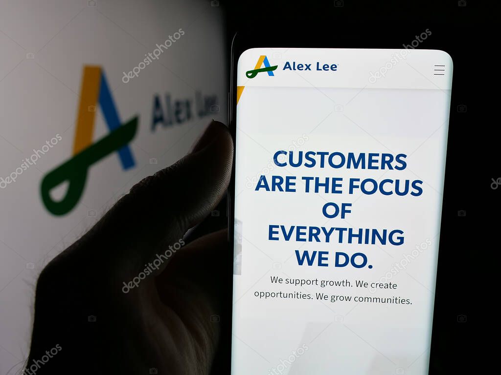 Stuttgart, Germany - 06-05-2025: Person holding cellphone with webpage of US grocery wholesale company Alex Lee Inc. on screen in front of logo. Focus on center of phone display.