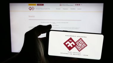 Stuttgart, Almanya - 07-28-2025: İnternet sayfasının önünde ekranda Letonya partisi Nacionala Apvieniba 'nın (NA) logosuna sahip cep telefonu tutan kişi. Telefon ekranına odaklan.