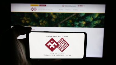 Stuttgart, Almanya - 07-28-2025: İnternet sayfası önünde ekranda Letonya partisi Nacionala Apvieniba 'nın (NA) logosuna sahip cep telefonu tutan kişi. Telefon ekranına odaklan.