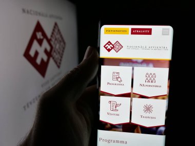 Stuttgart, Almanya - 07-28-2025: Letonya partisi Nacionala Apvieniba 'nın (NA) web sayfası logonun önünde duran kişi. Telefon ekranının merkezine odaklan.