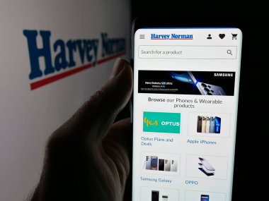 Stuttgart, Almanya - 08-25-2025: Avustralyalı perakende şirketi Harvey Norman 'ın web sayfası olan akıllı telefon sahibi kişi logonun önünde ekranda. Telefon ekranının merkezine odaklan.
