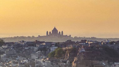 Arkasında büyük bir bina olan bir şehir manzarası. Gökyüzü turuncu ve mavinin bir karışımıdır. Sıcak ve huzurlu bir atmosfer yaratır.