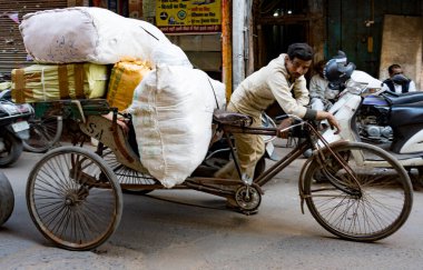 Çalışkan işçi, kalabalık bir caddede ağır yüklerle yüklü üç tekerlekli bisiklet pedalına basıyor.