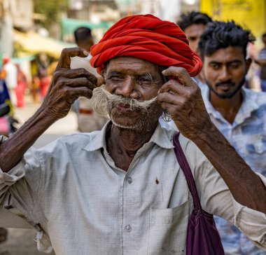 Kıdemli, kırmızı sarıklı Hintli adam, kalabalık bir sokak sahnesinde bıyığını düzeltiyor.