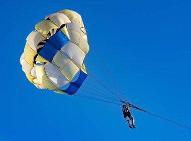 Güneşli bir günde, açık mavi gökyüzüne paraşütle atlayan kişi, sporun heyecanını gözler önüne seriyor.