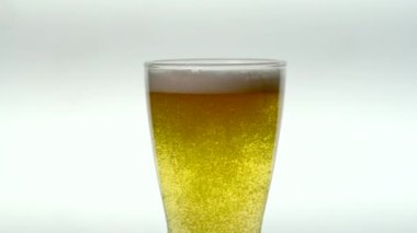 Köpük altında kabarcıklar yükselen soluk bira dolu bardak beyaz bir arka planda yavaşça dönüyor