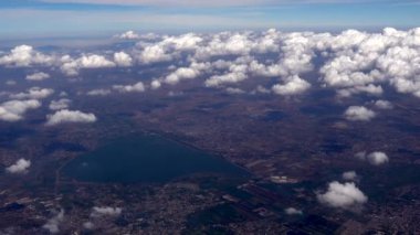 Meksiko şehrinin üzerinde uçan bir uçak ve bir göl, parlak mavi gökyüzünde dağınık bulutlar sergiliyor.